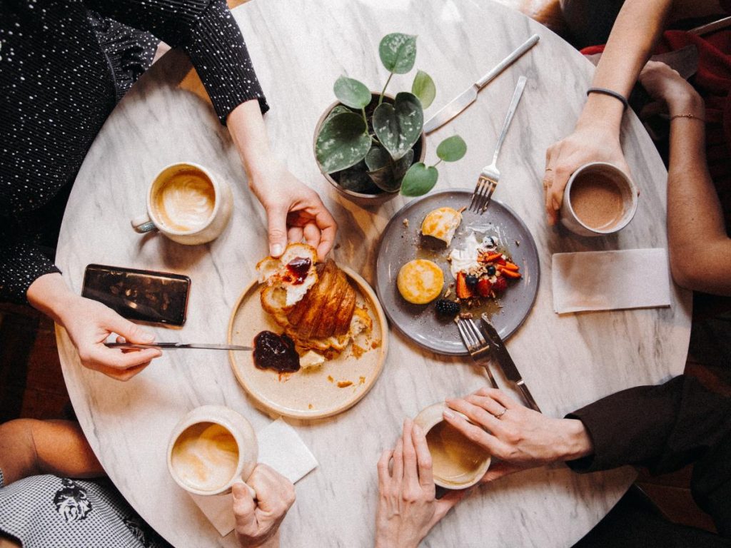 Quattro persone sorseggiano caffè e pasticcini attorno a un tavolo rotondo in marmo. L'atmosfera è accogliente e rilassata, con un centrotavola di piante e piatti da condividere.