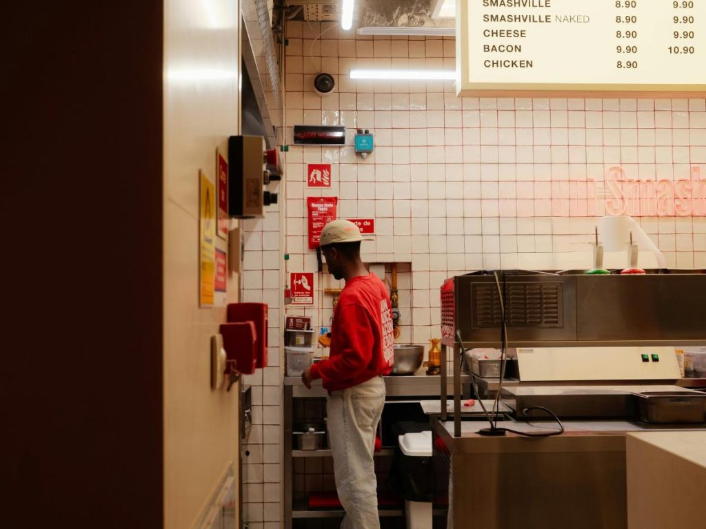 Una persona con una felpa rossa e un berretto bianco lavora in una cucina con pareti piastrellate. Sopra è visibile un menu. L'ambiente è vivace e ordinato.