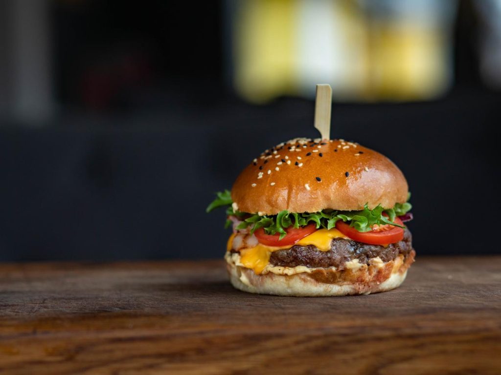 Un succulento cheeseburger su un tavolo di legno con un panino ai semi di sesamo, lattuga, pomodoro e formaggio fuso. Uno stuzzicadenti lo tiene insieme. Lo sfondo è sfocato.