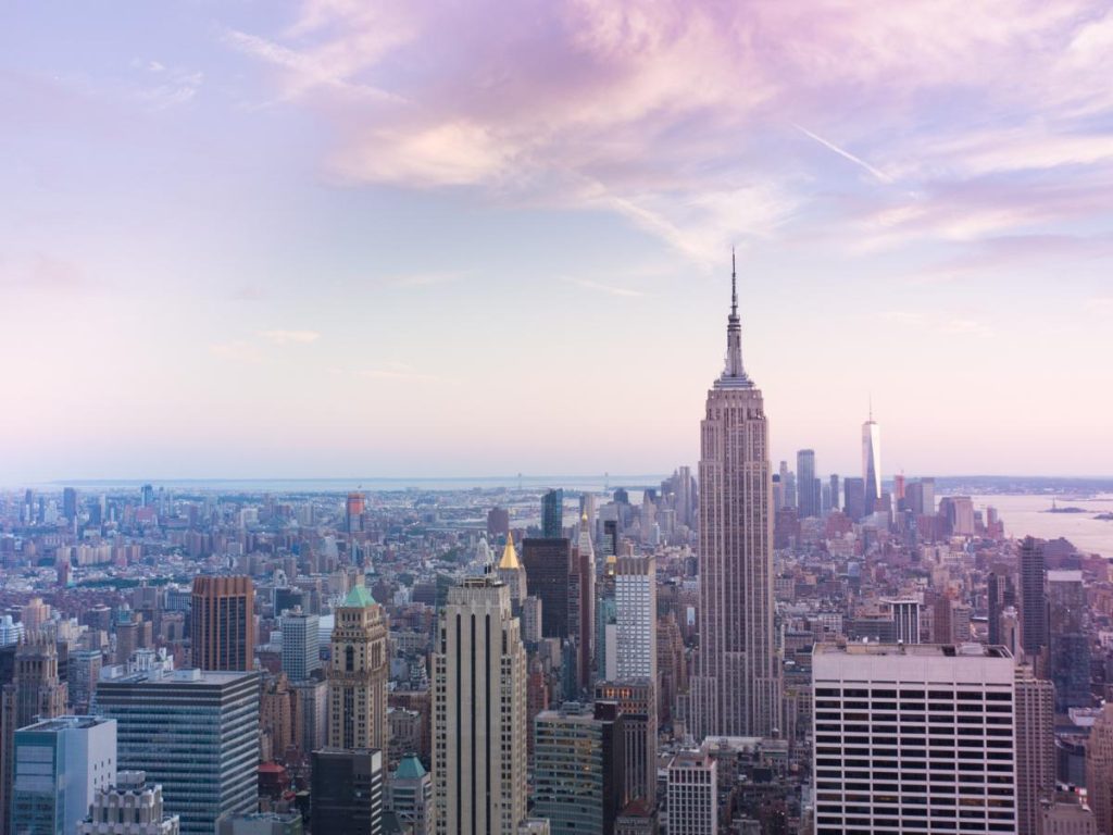 Vista aerea dello skyline di New York City al tramonto, con i grattacieli sotto un cielo rosa pastello e azzurro, che evoca un'atmosfera calma e vibrante.