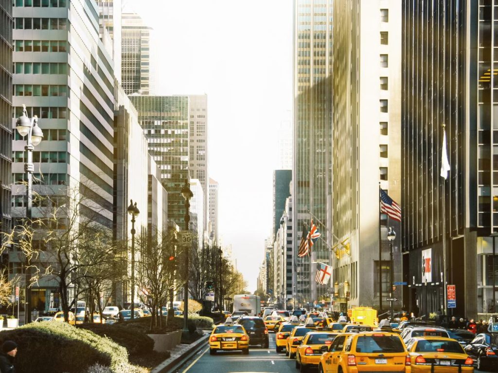 Una strada cittadina trafficata con alti grattacieli e numerosi taxi gialli nel traffico. Bandiere appese agli edifici e la scena è illuminata da una luce solare calda e intensa.