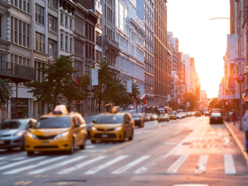Una vivace strada cittadina al tramonto, con taxi gialli e auto in movimento. Alti edifici costeggiano la strada e la calda luce del sole crea un'atmosfera serena e urbana.