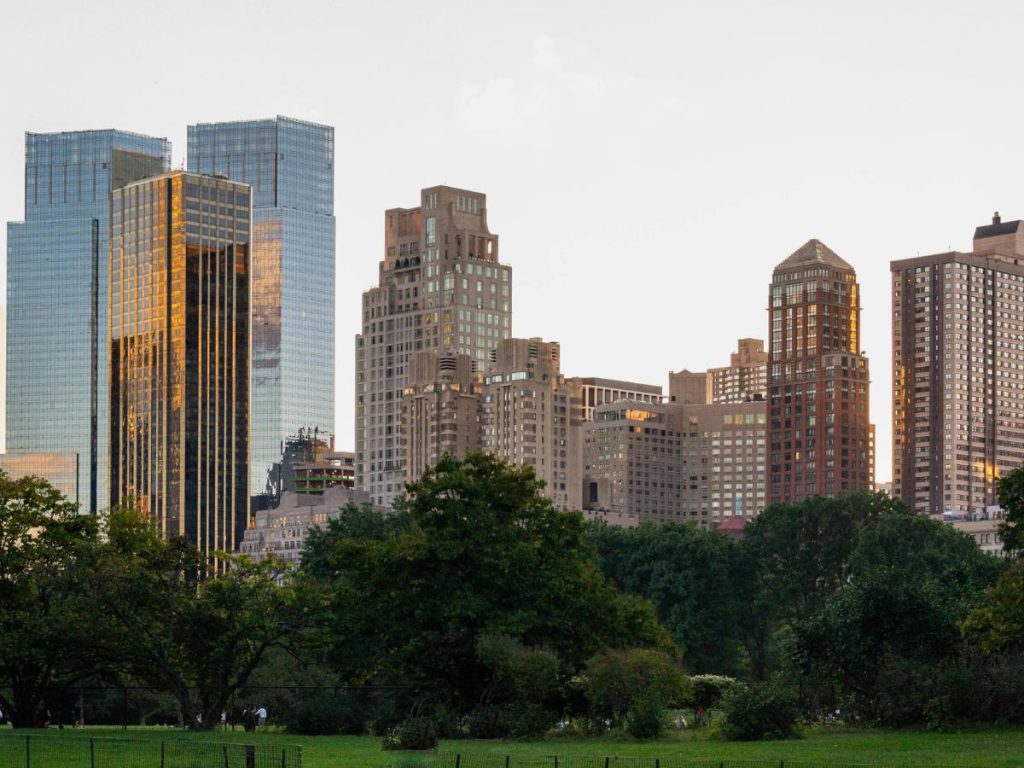 Lo skyline della città, con i suoi moderni grattacieli che riflettono un tramonto dorato, è circondato da rigogliosi alberi verdi. Lo scenario è tranquillo e pittoresco.