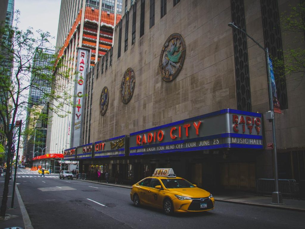 Un taxi giallo passa davanti all'iconico Radio City Music Hall di New York. Il cartellone pubblicitario mostra un annuncio dello spettacolo, con alti edifici che fiancheggiano entrambi i lati.