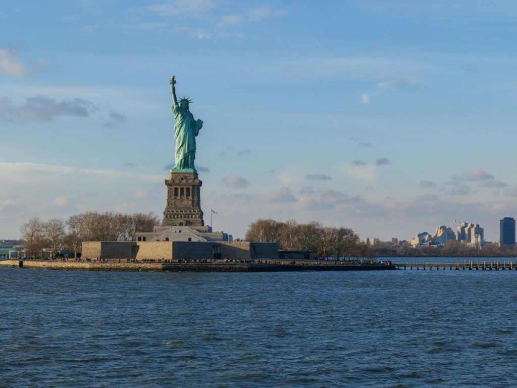La Statua della Libertà si erge su Liberty Island sotto un cielo azzurro. L'isola è circondata dall'acqua, con lo skyline di New York visibile in lontananza.