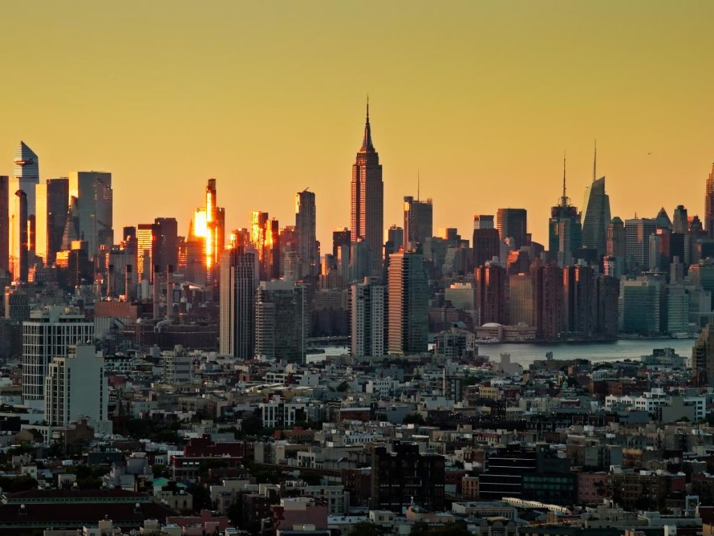 Lo skyline di New York City al tramonto, con gli edifici che si stagliano contro un cielo arancione, tra cui l'Empire State Building, trasmettendo un'atmosfera calma e maestosa.