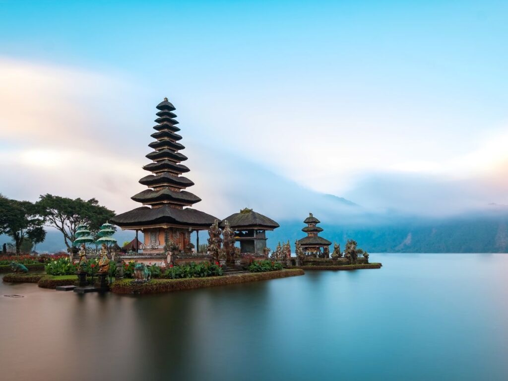 Un sereno tempio in Indonesia, tra le destinazioni da abbinare alle Maldive, in riva al lago con tetti a pagoda a più livelli che si riflettono nelle acque calme. Le montagne nebbiose e un'alba vibrante creano un'atmosfera di pace.