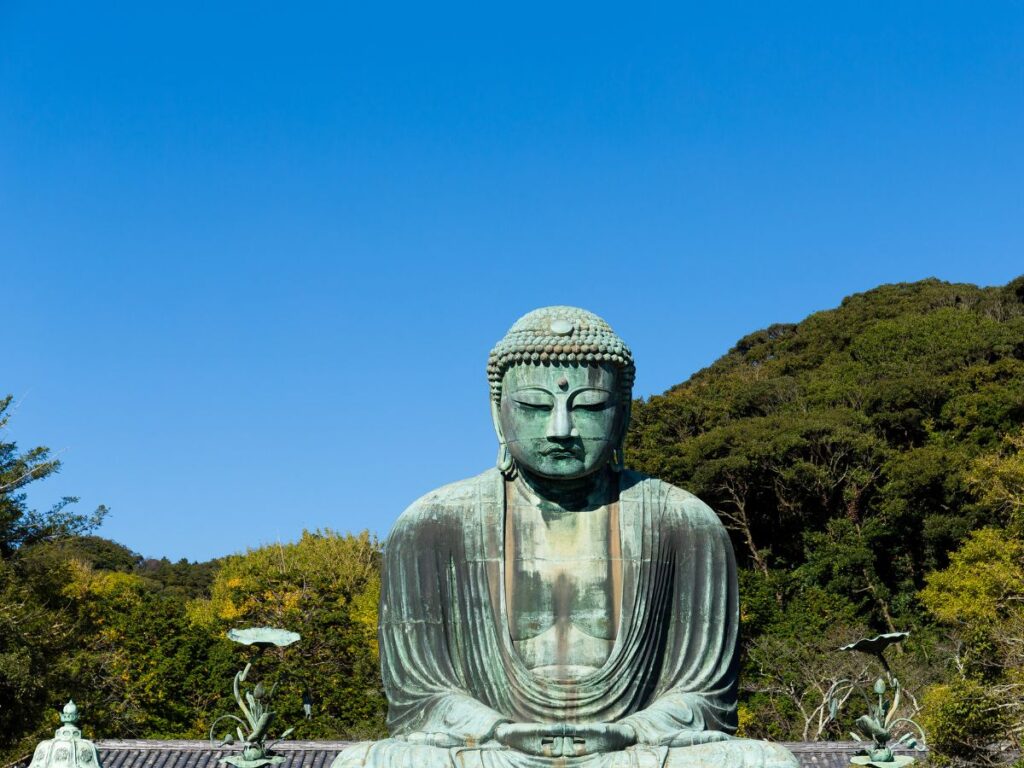Il Grande Buddha di Kamakura, una grande statua in bronzo, è collocato su uno sfondo di rigogliosi alberi verdi e un cielo azzurro intenso, evocando tranquillità.