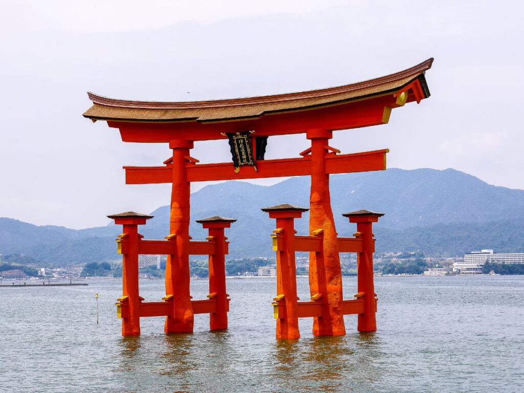 L'iconico torii rosso dell'isola sacra di Miyajima da vedere durante un viaggio in Giappone si erge nell'acqua, sullo sfondo di montagne e cielo nuvoloso, trasmettendo un senso di serenità e di patrimonio culturale.