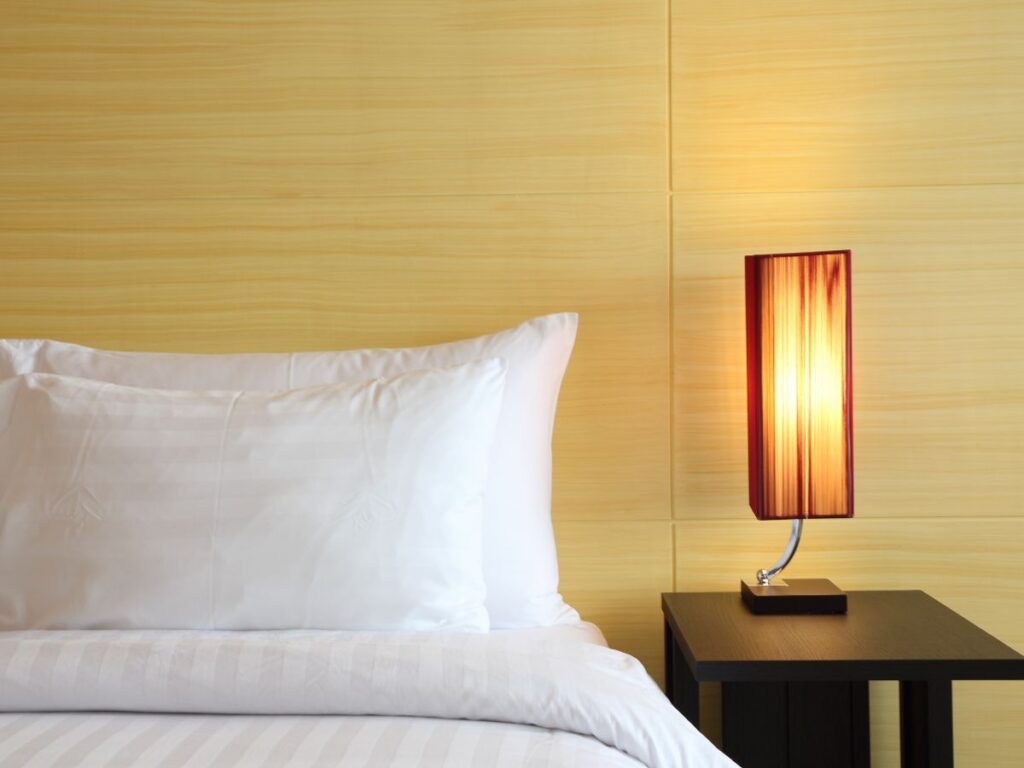 Un'accogliente scena da camera da letto con un cuscino bianco e un piumone sulla sinistra, accanto a un comodino in legno scuro con una lampada rettangolare alta e illuminata. Toni caldi di un boutique hotel dove dormire alle Maldive