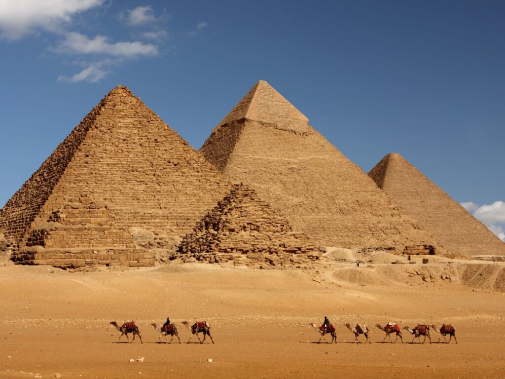 Tra i viaggi più belli da fare in Africa c'è l'Egitto. Scena desertica con tre antiche piramidi sotto un cielo azzurro e terso. Una fila di cammelli con cavalieri attraversa il primo piano sabbioso, evocando un viaggio senza tempo.