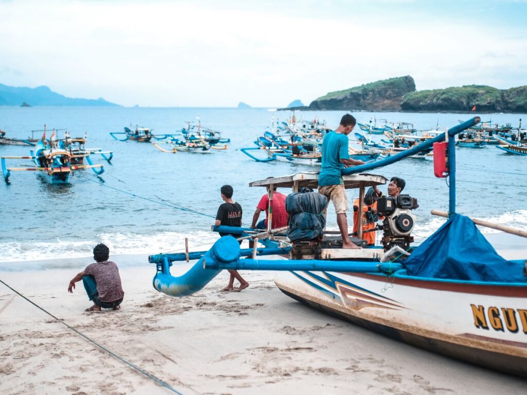 I pescatori preparano le barche su una spiaggia sabbiosa, con sullo sfondo diverse imbarcazioni colorate che solcano il mare calmo, sotto un cielo azzurro e nuvoloso. La scena è serena e animata.