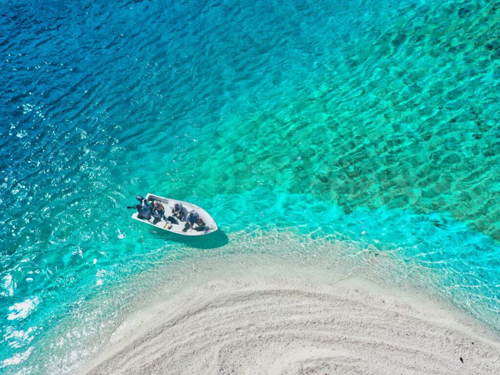 Vista aerea di una piccola barca con persone su acque turchesi vicino a una spiaggia sabbiosa. Il gradiente dell'acqua vira dal blu intenso al verde acqua, evocando un'atmosfera serena.