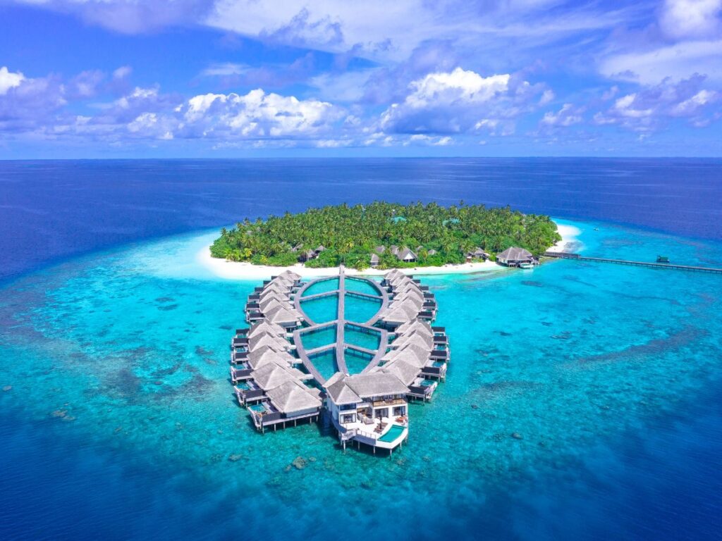 Vista aerea di un'isola tropicale circondata da un oceano cristallino, con ville sull'acqua disposte a semicerchio dove dormire alle Maldive. Una vegetazione lussureggiante ricopre l'isola.