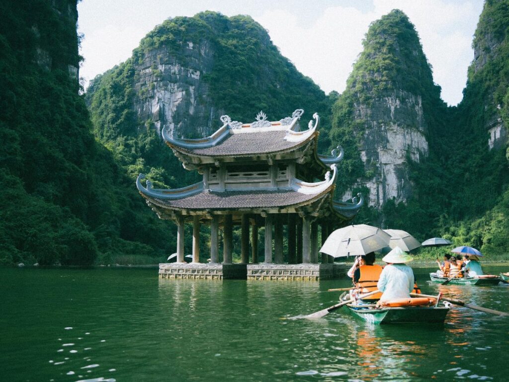 Barche con turisti su un lago circondato da lussureggianti montagne verdi, che si avvicinano a una pagoda tradizionale con tetto decorato, creando un'atmosfera serena.