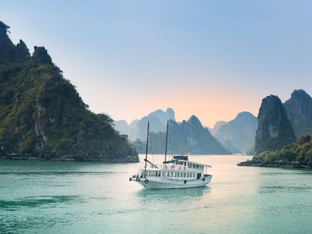 Una tranquilla barca bianca naviga attraverso acque turchesi, circondata da lussureggianti isolotti rocciosi, sotto un cielo mite al tramonto, evocando tranquillità ed esplorazione durante un viaggio di 12 giorni in Vietnam