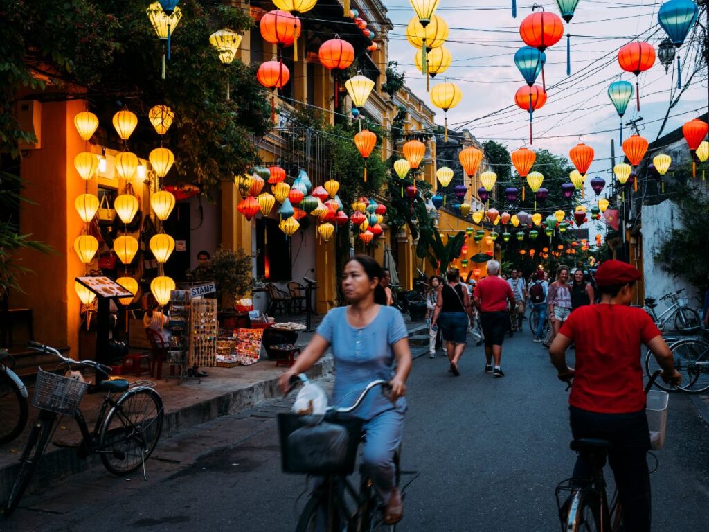 Una vivace scena di strada al tramonto, con lanterne colorate in cielo. La gente cammina e va in bicicletta lungo la stretta strada, con negozi e alberi lungo i lati, creando un'atmosfera vivace e festosa.