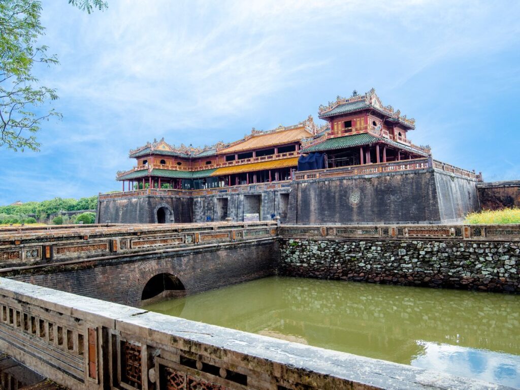 Antico palazzo vietnamita con tetti decorati in rosso e oro, circondato da mura di pietra. Un fossato riflette il cielo azzurro e terso, evocando un'atmosfera storica. Da visitare durante un viaggio di 12 giorni in Vietnam