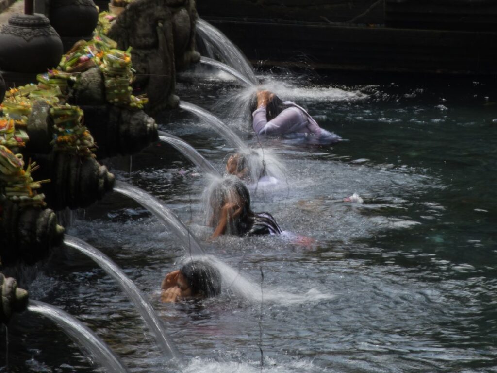 Le persone si immergono in una tranquilla piscina del tempio sotto getti di pietra, adornati con offerte colorate, che trasmettono tranquillità e purificazione spirituale.