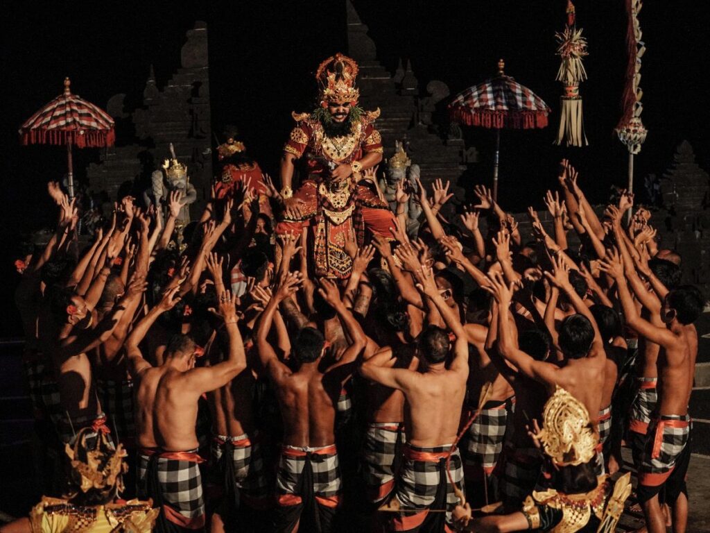 Una vivace scena di danza tradizionale notturna con un artista centrale in costume elaborato circondato da ballerini a torso nudo che stendono le braccia verso l'alto, trasmettendo unità ed energia.