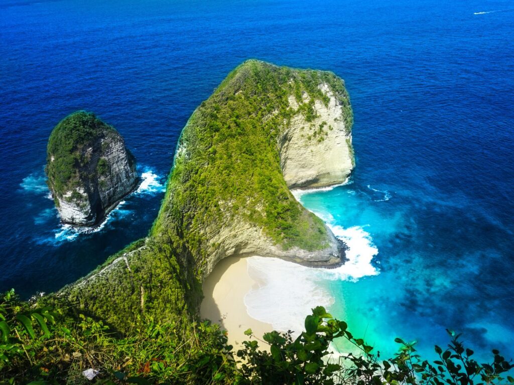 Vista aerea di una lussureggiante scogliera verde che si protende nell'oceano blu intenso, circondando un'appartata spiaggia di sabbia bianca. Il paesaggio trasmette tranquillità e bellezza naturale.