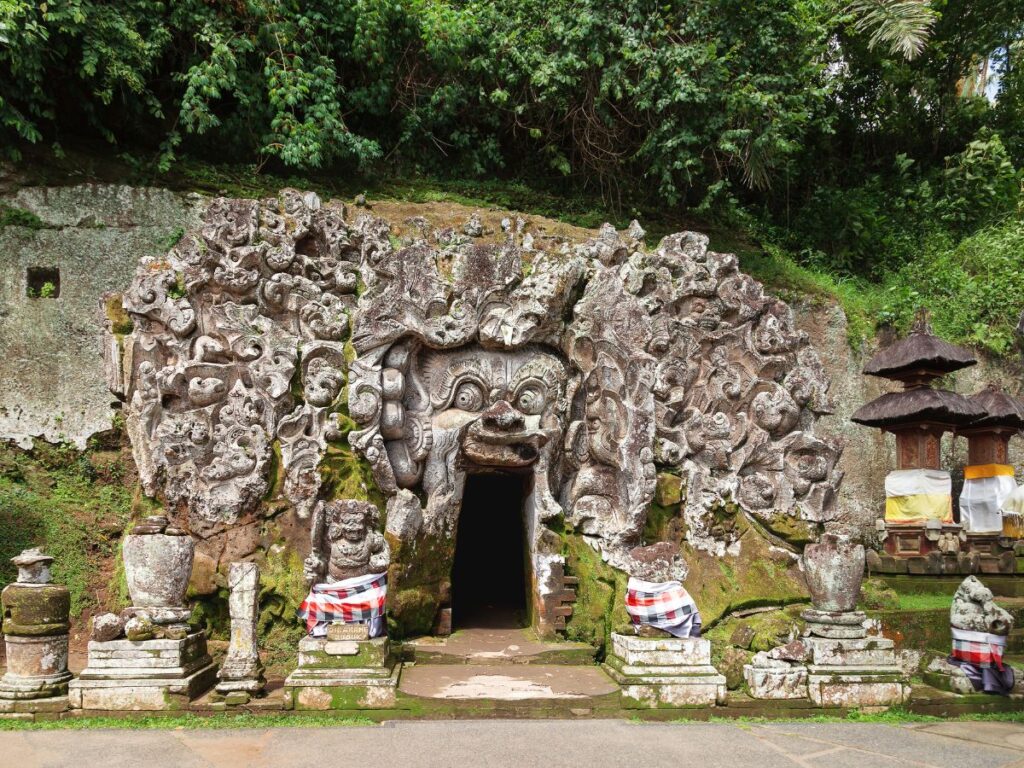 Ingresso di una grotta in pietra con intricate incisioni di volti e figure, circondato da una vegetazione lussureggiante. Alcune statue sono ricoperte da tessuti a scacchi, che conferiscono un'atmosfera mistica.
