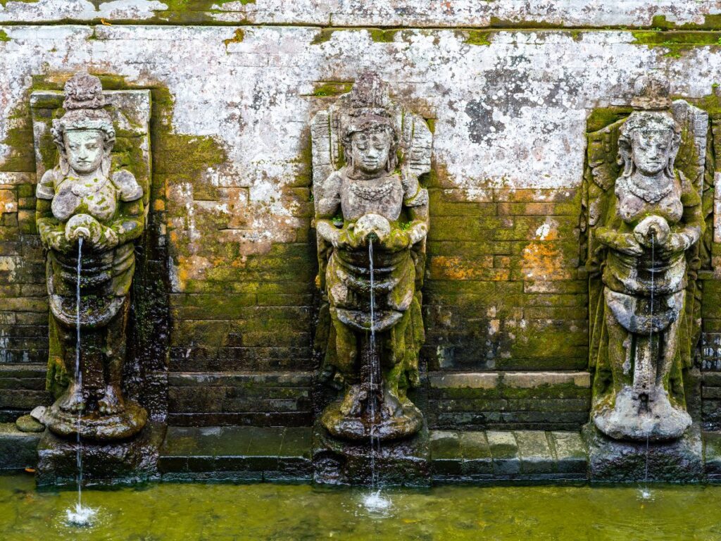 Tre antiche statue di pietra con copricapi e dettagli intricati si ergono contro un muro consumato dal tempo. L'acqua scorre da ogni statua in una piscina verde sottostante.