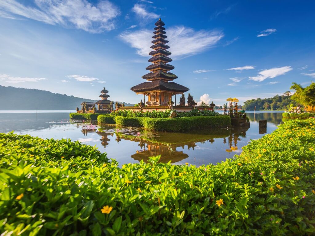 Paesaggio sereno con un tempio balinese con tetti a più livelli in mezzo a un lago calmo, circondato da una vegetazione lussureggiante e fiori gialli sotto un cielo azzurro e terso a Bali e Nusa Penida