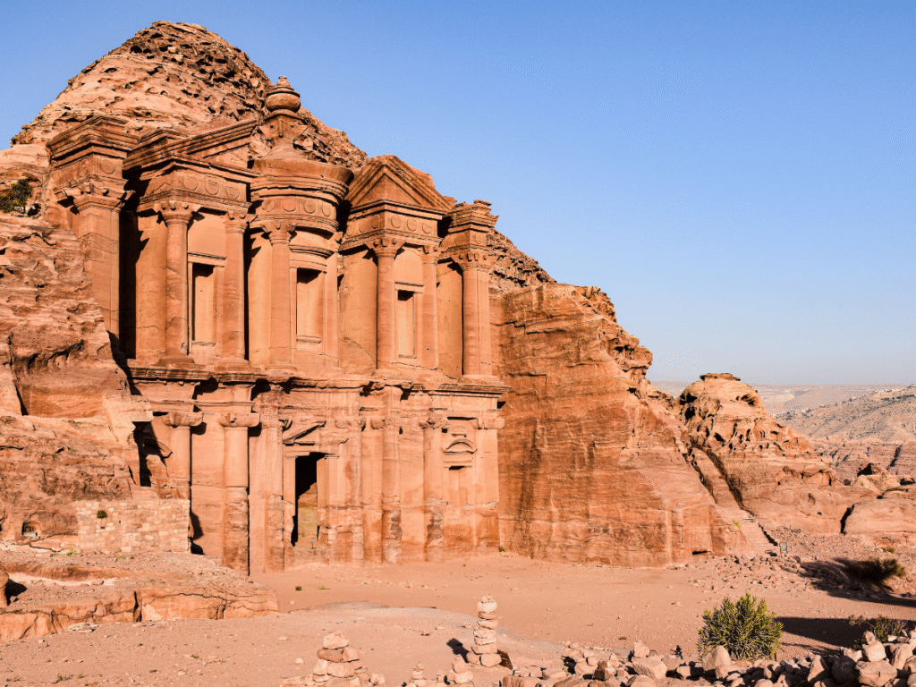 Antica facciata in pietra di un monastero a Petra, in Giordania, scavata nella roccia rossa. Il cielo azzurro e il paesaggio desertico evocano un senso di stupore e storia.