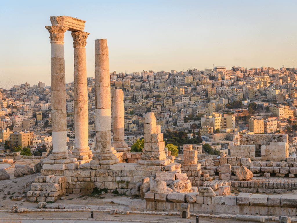 Antiche colonne di pietra di un tempio in rovina si ergono sotto un tramonto dorato, con un vasto paesaggio urbano pieno di edifici beige densamente ammassati sullo sfondo.
