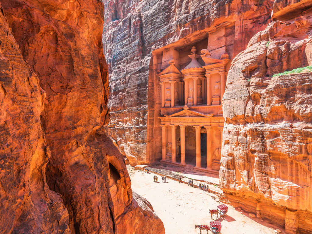 L'immagine mostra l'antica facciata scavata nella roccia del Tesoro di Petra, in Giordania, circondata da imponenti scogliere rosso-arancio. Persone e cammelli in piedi sotto, ne esaltano la grandiosità.