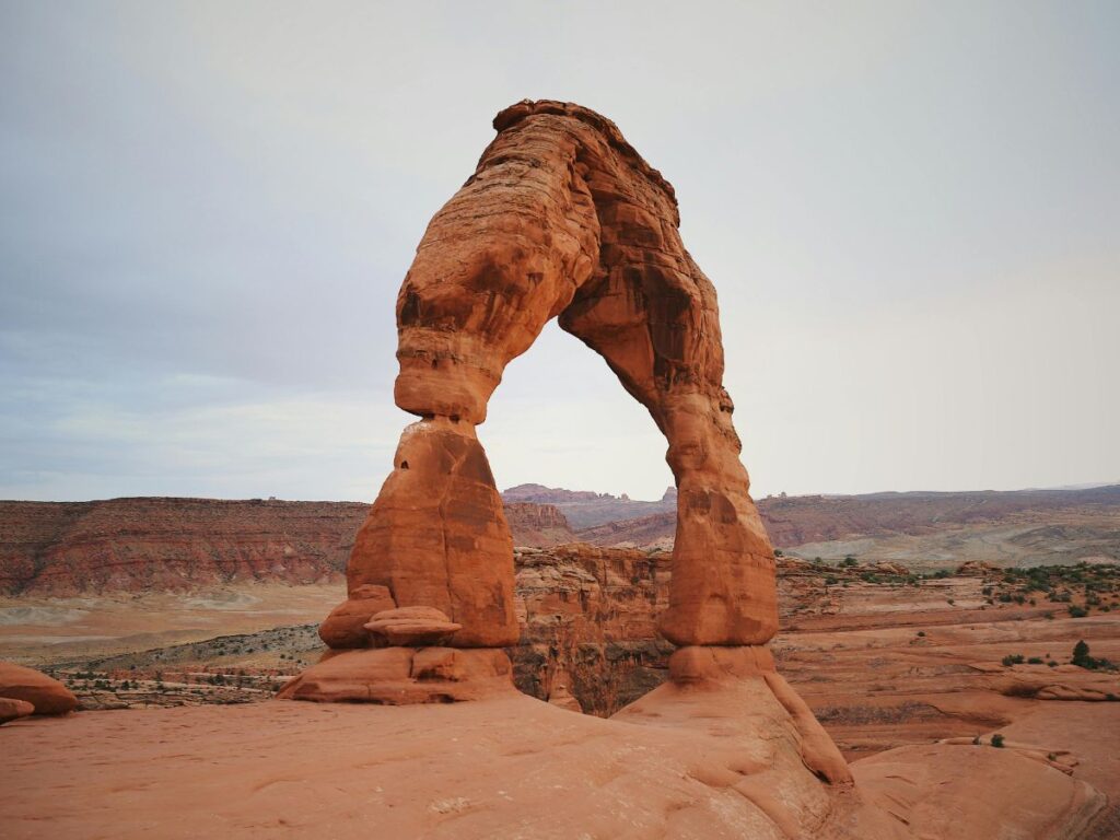 Un grande arco naturale di roccia rossa si erge imponente sotto un cielo pallido in un paesaggio desertico, creando un'atmosfera serena e maestosa.