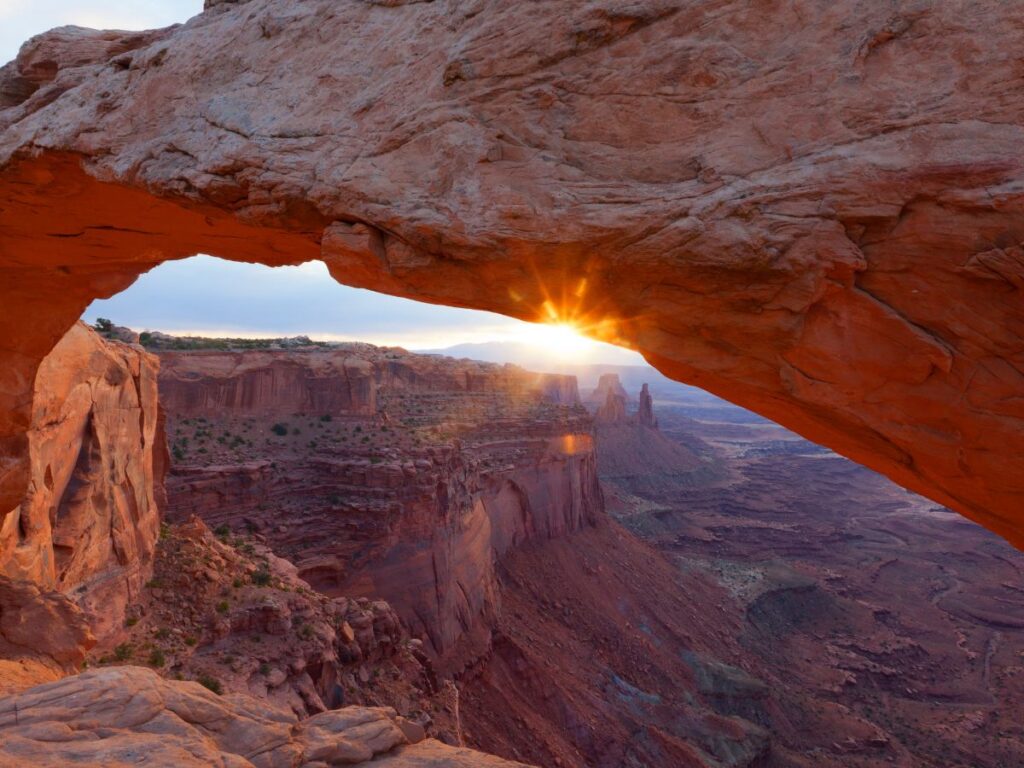 Alba spettacolare vista attraverso un arco di roccia naturale, che mette in risalto le vivaci tonalità arancioni. Sotto, il vasto e aspro paesaggio del canyon si estende sotto un cielo terso.