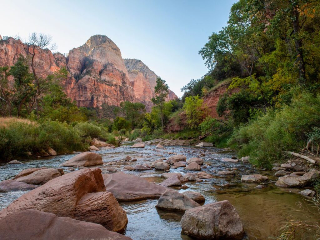 Uno scenario sereno di un fiume roccioso che scorre attraverso un canyon verdeggiante e lussureggiante. Sullo sfondo si ergono aspre scogliere sotto un cielo azzurro e terso, creando un'atmosfera di pace.