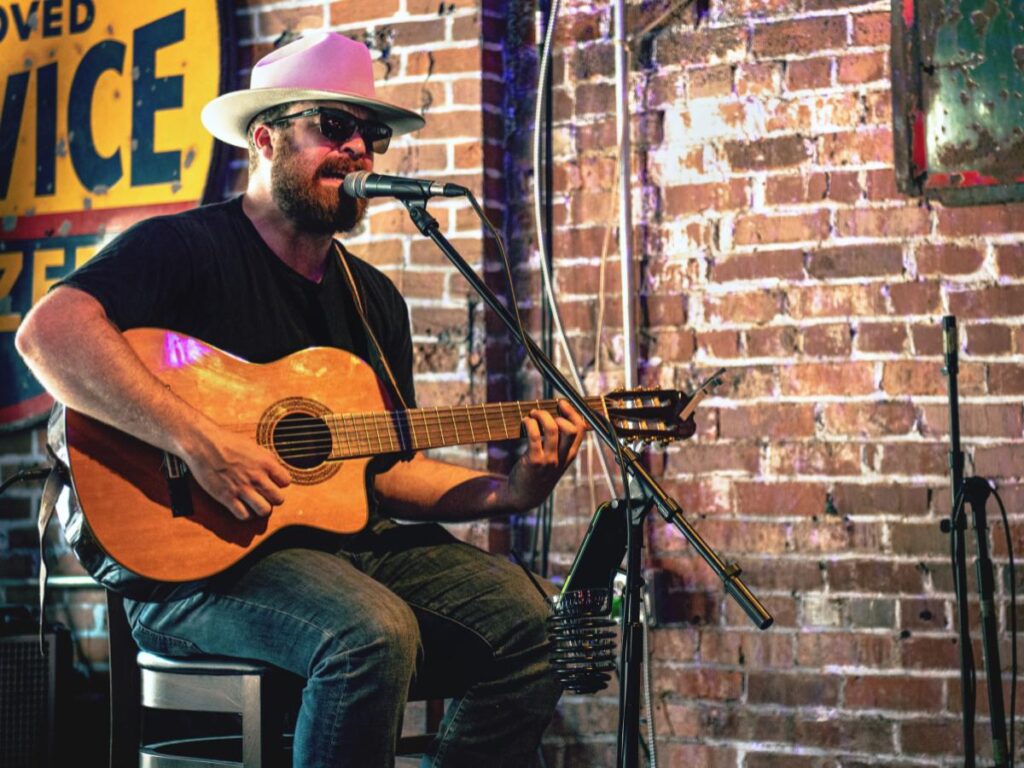 Un uomo con un cappello bianco e occhiali da sole suona una chitarra acustica e canta al microfono. È seduto contro un muro di mattoni, emanando un'atmosfera rilassata e intima.