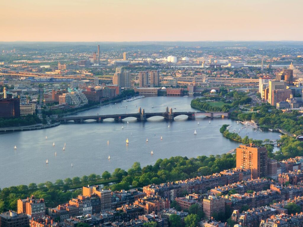 Vista aerea di Boston al tramonto, con il fiume Charles, barche a vela, rigogliosi alberi verdi e storici edifici in mattoni, che emanano un'atmosfera calma e urbana.
