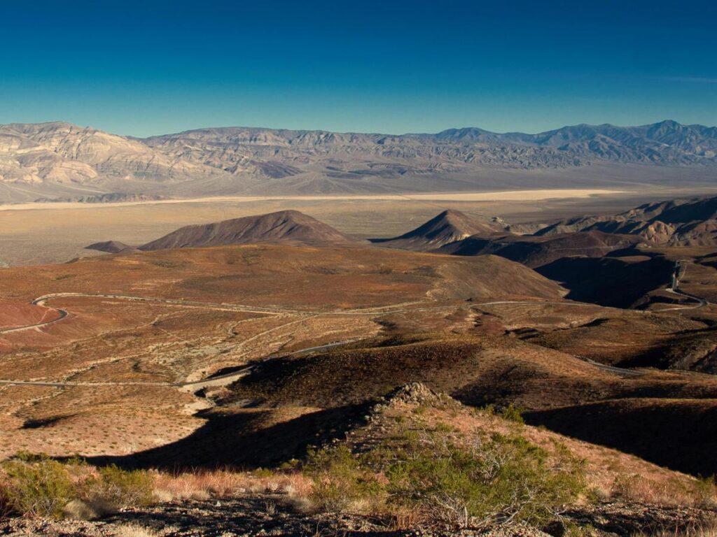 Paesaggio desertico con dolci colline, vegetazione rada e montagne aspre in lontananza sotto un cielo azzurro e terso, che trasmette un senso di vastità e tranquillità.