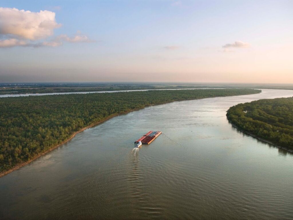 Vista aerea di una chiatta che naviga su un ampio fiume, circondata da fitte foreste verdi sotto un cielo azzurro e terso con qualche nuvola. Tranquillo e spazioso.