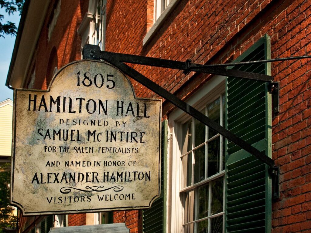 Insegna storica per Hamilton Hall, progettata da Samuel McIntire nel 1805, montata su un edificio in mattoni rossi con persiane verdi. Tonalità calda e invitante.