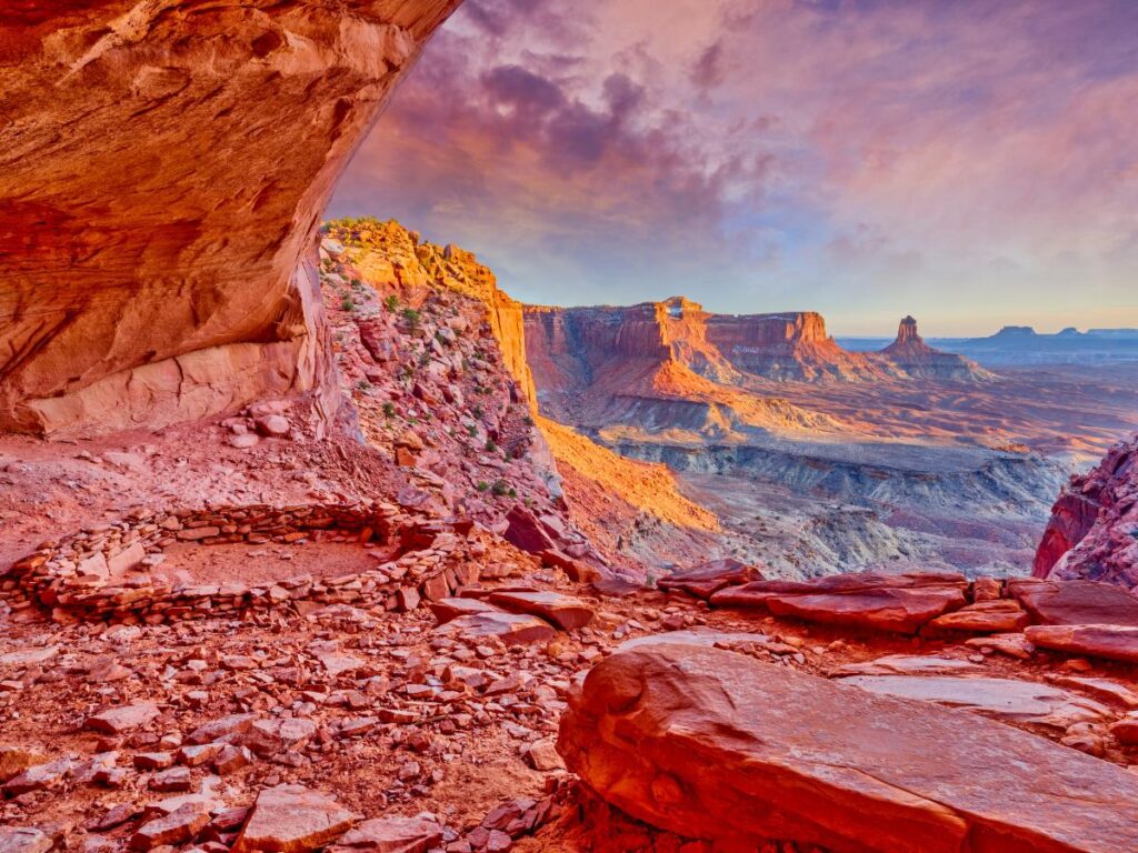 Vista da una sporgenza del canyon al tramonto, con formazioni rocciose rosse e un cerchio di pietre. Il cielo è vibrante di viola e arancioni, creando un'atmosfera serena.