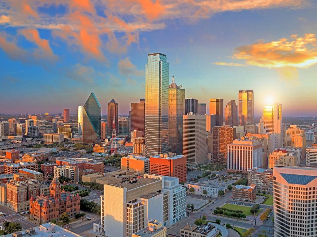 Lo skyline urbano vibrante di Dallas in Texas al tramonto, caratterizzato da alti grattacieli con vetrate riflettenti. La luce calda e dorata proietta un bagliore sereno, mettendo in risalto un cielo limpido e colorato.