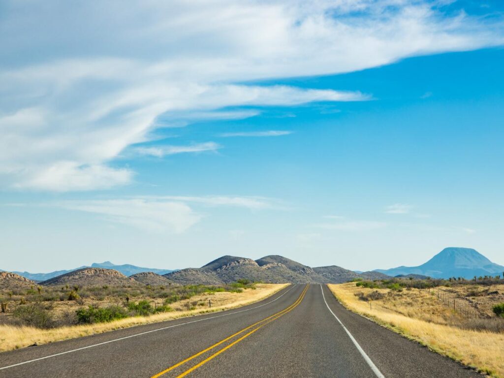 Una lunga strada dritta si snoda verso montagne lontane, sotto un ampio cielo azzurro con nuvole sparse. Erba secca e arbusti costeggiano la strada, trasmettendo solitudine.
