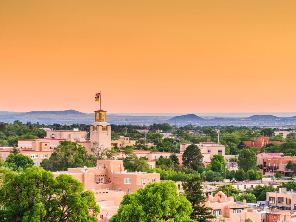 Vista aerea di Santa Fe, nel New Mexico, con edifici in terracotta, vegetazione lussureggiante e montagne in lontananza, sotto un caldo cielo arancione al tramonto. Atmosfera tranquilla e serena da scoprire durante un tour nell'ovest degli stati uniti