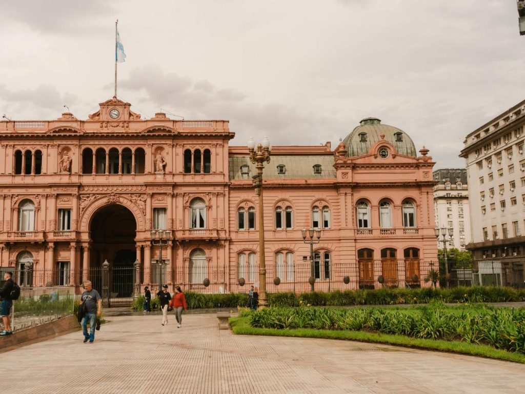 Veduta esterna della Casa Rosada a Buenos Aires, l'iconico palazzo presidenziale argentino dal caratteristico colore rosa, situato davanti a una piazza con persone che camminano.