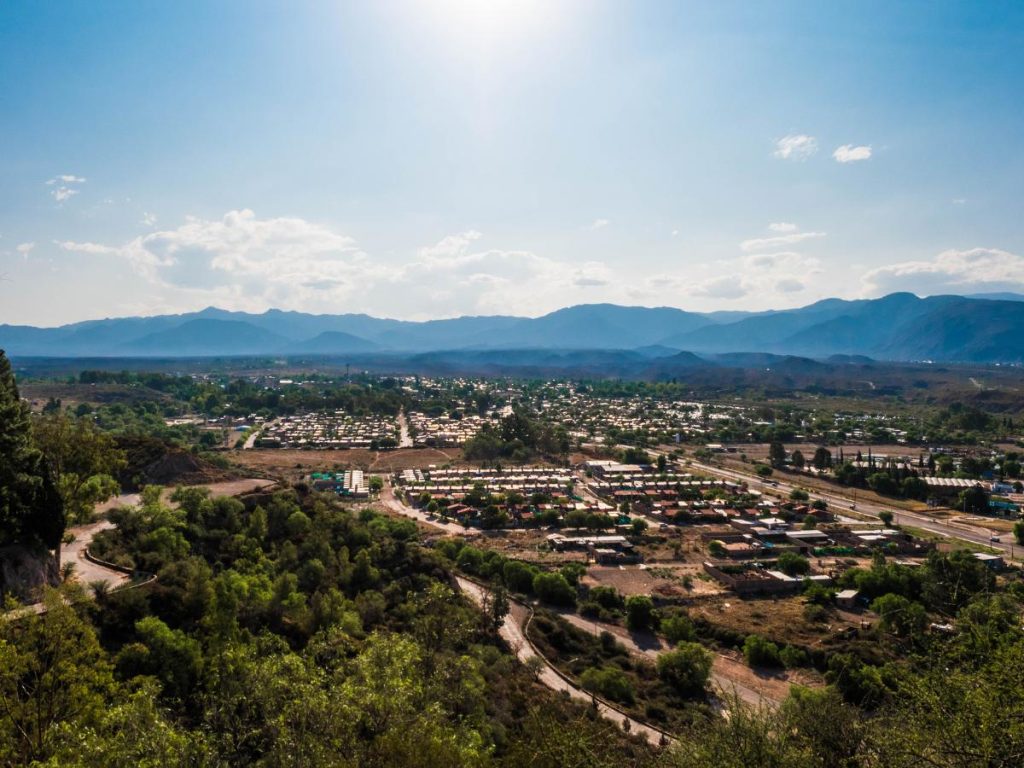 Una veduta panoramica dall'alto di una zona residenziale di Mendoza, circondata da vegetazione e con le maestose montagne delle Ande sullo sfondo sotto un cielo azzurro.