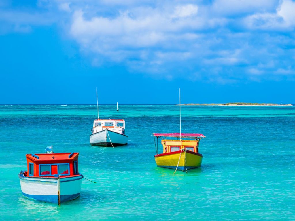 Tre piccole barche da pesca colorate (bianca, gialla e rossa) che galleggiano nelle calme acque turchesi dei Caraibi ad Aruba, sotto un cielo azzurro con nuvole bianche, tra le destinazioni perfette da visitare in estate