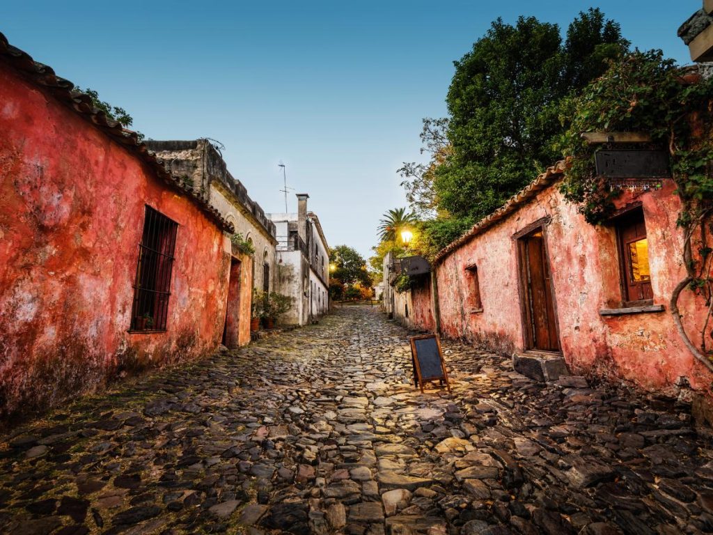 Una suggestiva strada acciottolata a Colonia del Sacramento, Uruguay, fiancheggiata da antiche case coloniali dalle pareti color pastello e alberi rigogliosi sotto un cielo crepuscolare.