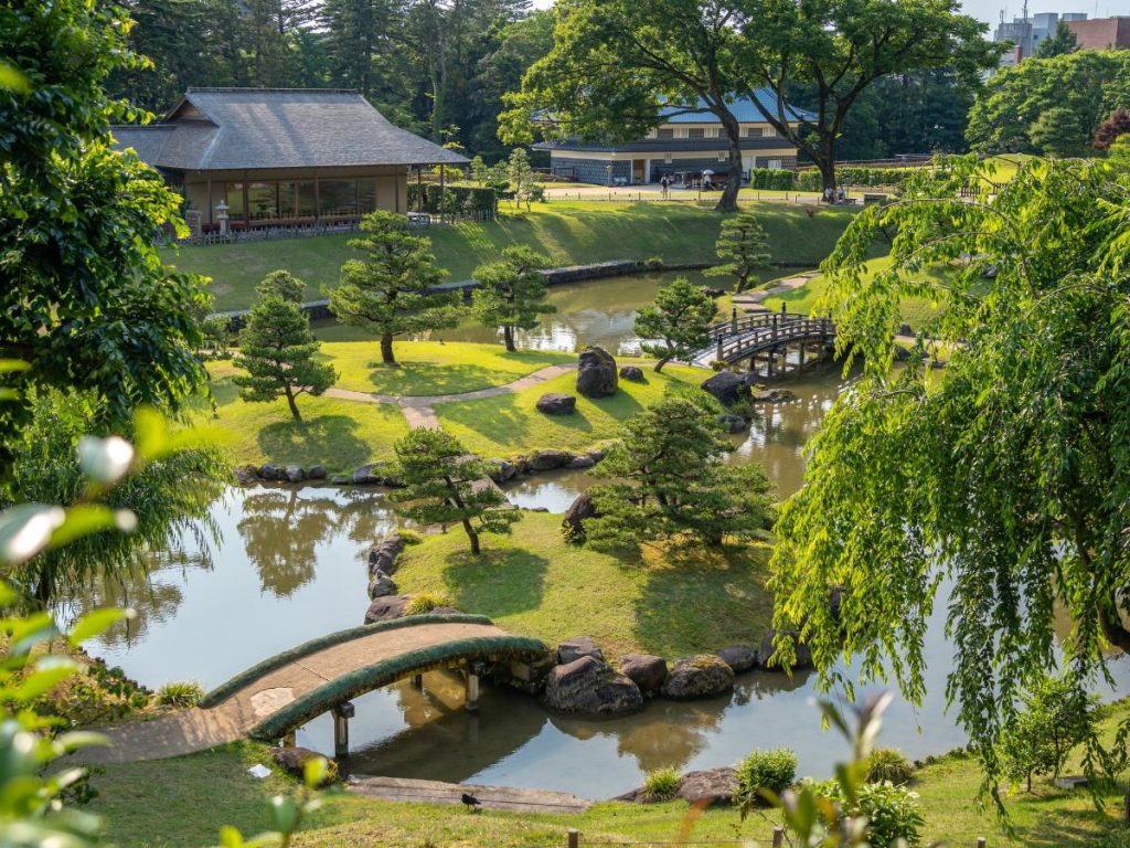 Un tradizionale giardino giapponese con un laghetto sereno, piccoli ponti in legno e pietra, alberi di pino curati e un edificio in stile classico (padiglione) immerso nel verde