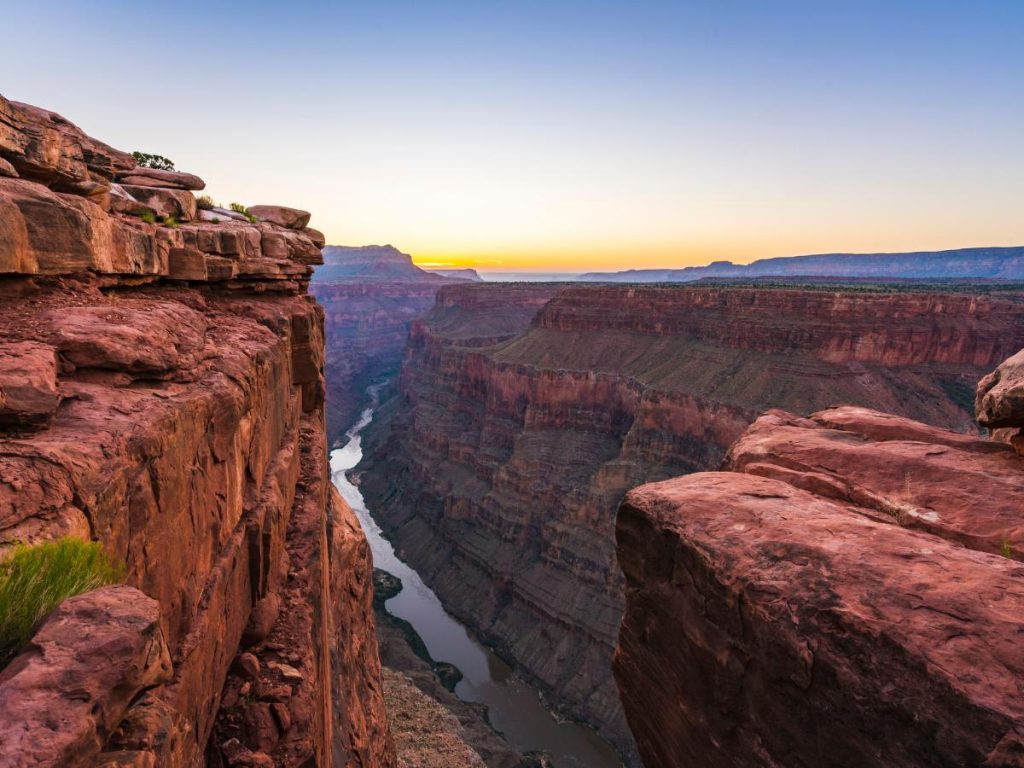 Una vista spettacolare del Grand Canyon al tramonto, con le stratificazioni rocciose rosse che scendono verso il fiume Colorado e un cielo sfumato di arancione e blu