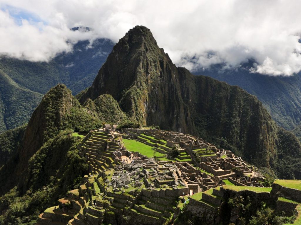 Veduta aerea delle antiche rovine inca di Machu Picchu in Perù, con le caratteristiche terrazze agricole verdi, le strutture in pietra e le imponenti vette montuose circondate dalle nuvole